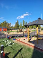 Kuvassa päiväkodin piha-aluetta, edessä iso hiekkalaatikko katoksella. Taaempana näkyy keinuja, asfalttinen pyöräilyrata, nurmi aluetta sekä pienten lasten leikkivälineitä